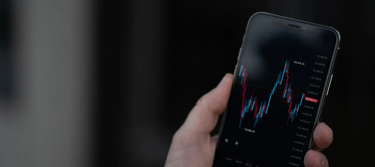 essoa segurando um smartphone com gráfico de candlestick exibido na tela, representando análise de mercado financeiro ou de criptomoedas em tempo real.