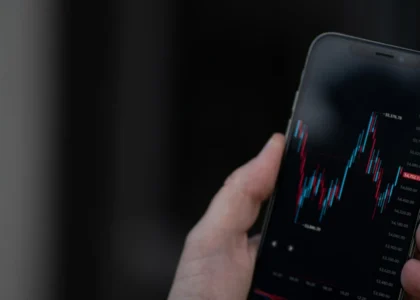 essoa segurando um smartphone com gráfico de candlestick exibido na tela, representando análise de mercado financeiro ou de criptomoedas em tempo real.