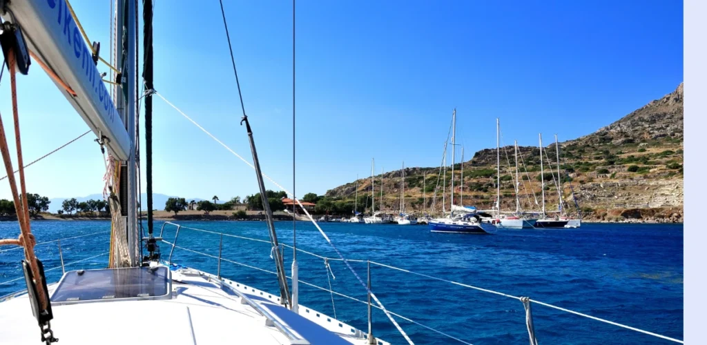 Imagem capturada da lateral de um veleiro de luxo, mostrando o convés branco e águas azuis cristalinas, com montanhas e outras embarcações ao fundo. Representa o potencial turístico e de aluguel do ativo tokenizado.