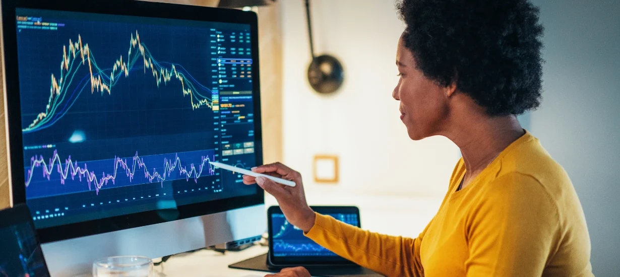 Mulher analisando gráficos financeiros em uma tela de computador, comparando plataformas de negociação como Deribit e GMX, com indicadores técnicos visíveis.