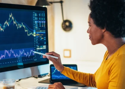 Mulher analisando gráficos financeiros em uma tela de computador, comparando plataformas de negociação como Deribit e GMX, com indicadores técnicos visíveis.